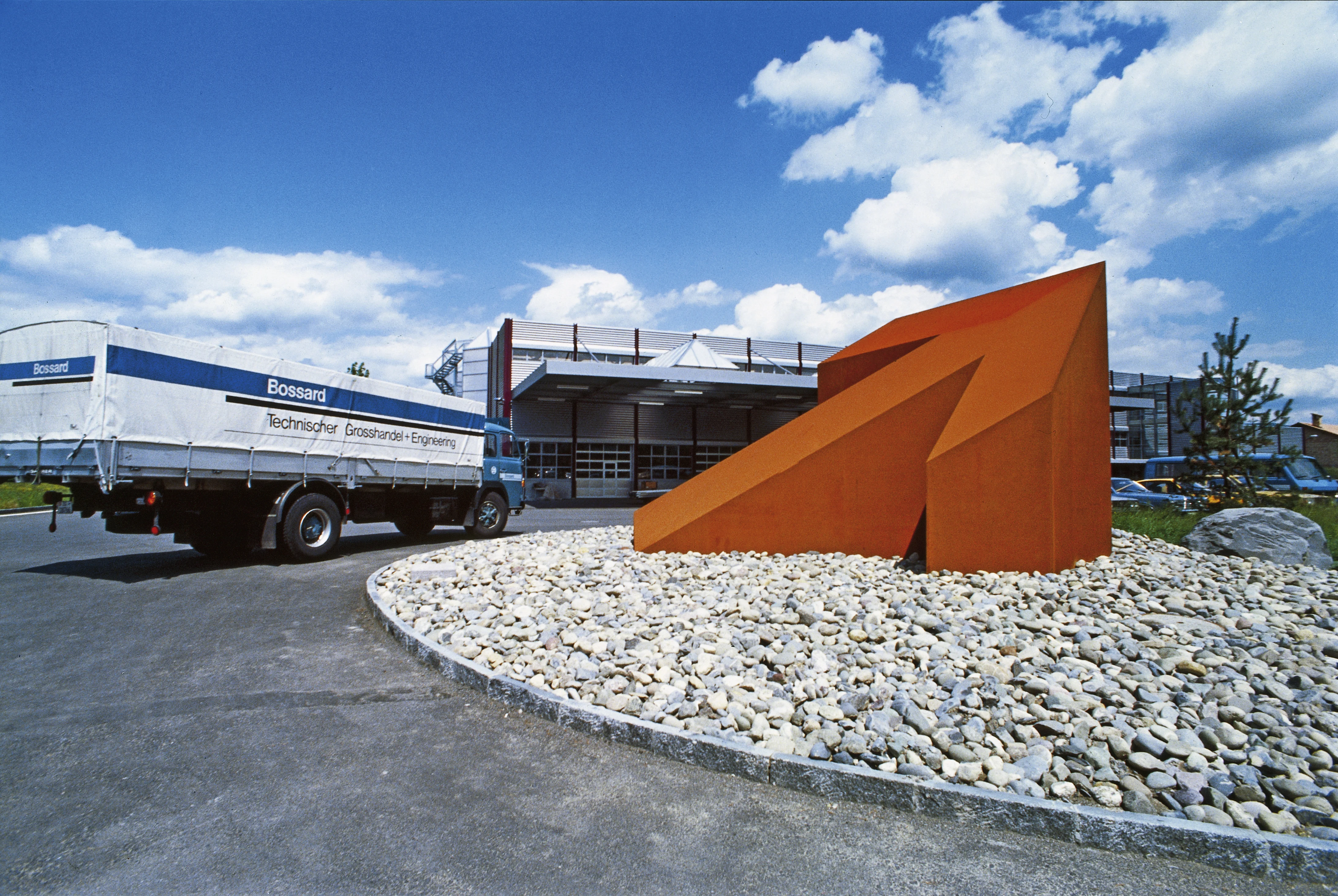 Historical image of a large orange arrow sculpture in a roundabout, with a Bossard truck and warehouse in the background.